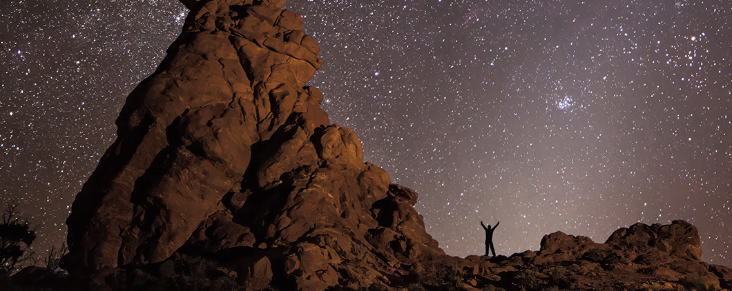 Photo of a person standing next to a desert rock formation at night with endless stars in sky